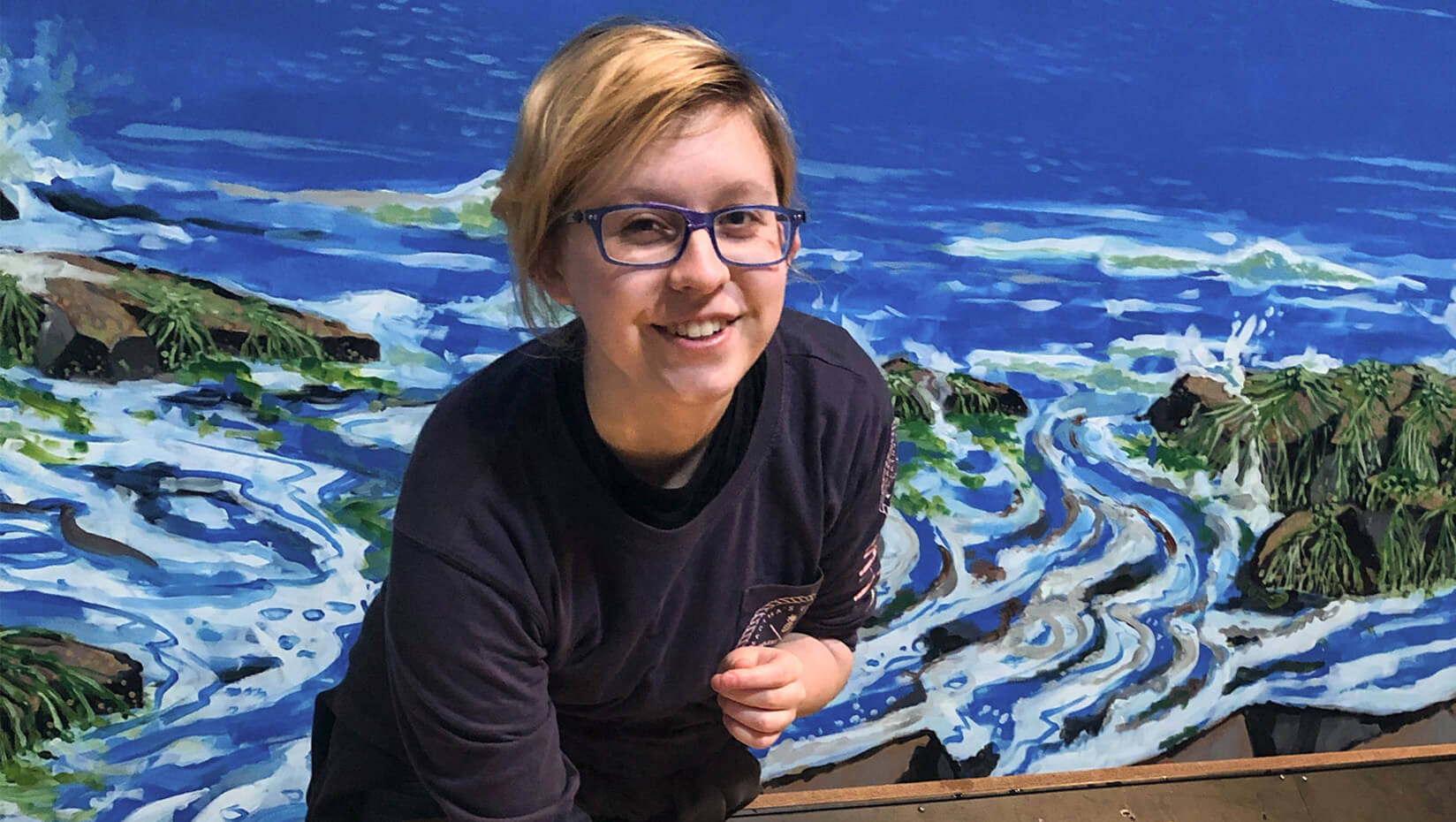 An image of Brynn Yarbrough standing in front of a painting of the ocean.